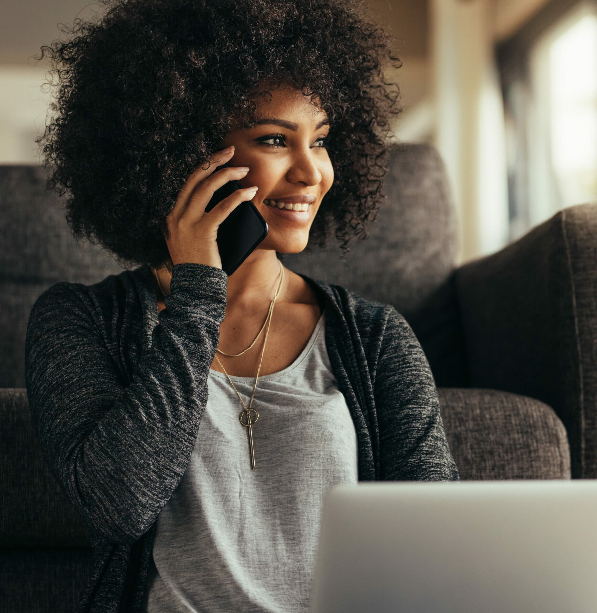 A young woman at home with a laptop talking on cell phone and smiling.